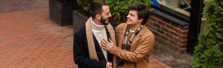 trendiger schwuler Mann im beigen Mantel, der die Brust eines bärtigen Freundes berührt, während er sich im Freien anlächelt, Banner