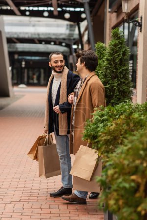 Foto de Socios gay de moda con bolsas de compras sonriendo el uno al otro cerca de plantas verdes en la calle - Imagen libre de derechos