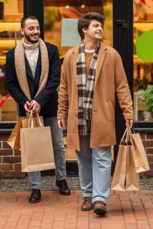 longitud completa de moda y feliz gay los hombres con compras bolsas cerca borrosa escaparate en la calle urbana