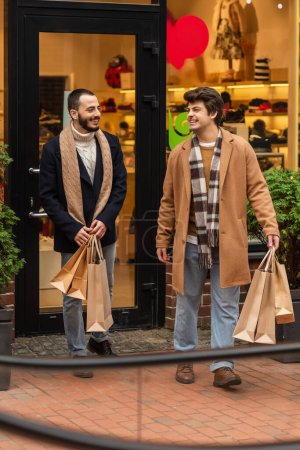 full length of cheerful gay men in stylish clothes holding shopping bags near store on city street