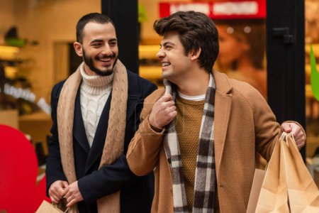 Foto de Sonriente gay hombre celebración de compras bolsas y ajuste bufanda cerca de moda barbudo novio y borrosa escaparate - Imagen libre de derechos