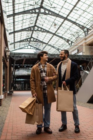 Foto de Longitud completa de socios gay con bolsas de compras sonriendo el uno al otro en la calle bajo techo transparente - Imagen libre de derechos