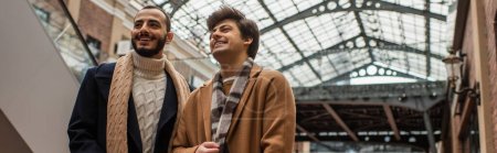 Téléchargez les photos : Jeune et à la mode gay les hommes souriant et regardant loin sous le toit transparent à l'extérieur, bannière - en image libre de droit