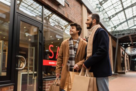 sonriente pareja gay con bolsas de compras mirando escaparate en la calle urbana