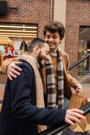 smiling man with shopping bags hugging bearded gay partner on escalator