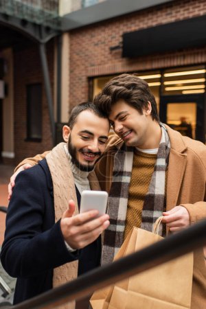 glücklicher schwuler Mann mit Einkaufstaschen umarmt trendigen bärtigen Freund, während er auf das Handy schaut