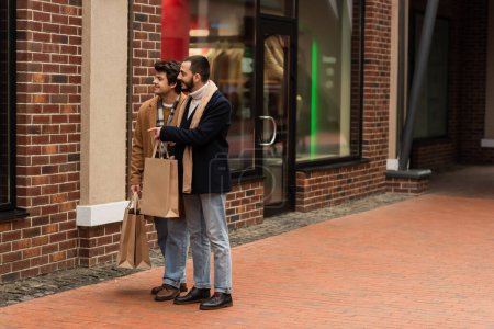 Vollbärtiger schwuler Mann mit Einkaufstüten zeigt auf Schaufenster in der Nähe eines jungen Freundes