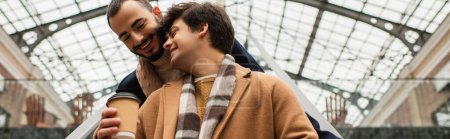 Téléchargez les photos : Faible angle de vue de gai gay homme tenant café pour aller près petit ami sous le toit transparent à l'extérieur, bannière - en image libre de droit