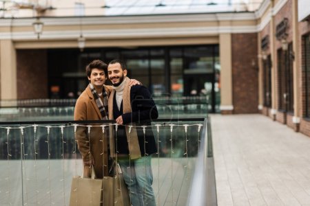 trendige und glückliche schwule Männer mit Einkaufstüten, die in der Nähe von Glaszaun und verschwommenem Gebäude stehen