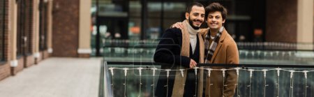 fashionable gay men in trendy coats looking away near glass fence and blurred building, banner