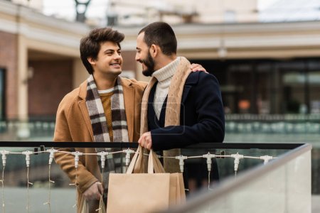 Photo for Happy gays in trendy coats and scarfs holding shopping bags and looking at each other on urban street - Royalty Free Image