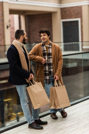 volle Länge der modischen Homosexuell Paar mit Einkaufstüten reden in der Nähe von Glaszaun auf der städtischen Straße