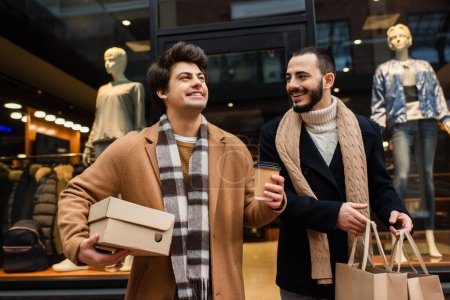 Foto de Barbudo gay hombre con compras bolsas mirando feliz novio con papel taza y zapatero cerca escaparate con maniquíes - Imagen libre de derechos