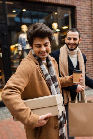 fröhlicher schwuler Mann blickt auf Schuhkarton neben lächelndem Freund mit Einkaufstasche und Pappbecher in verschwommener Vitrine 