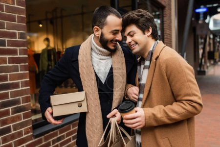glückliches schwules Paar mit Einkäufen und Pappbecher steht neben verschwommener Vitrine auf der Stadtstraße