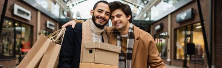 Foto de Alegre hombre con bolsas de compras abrazando pareja gay con cajas de zapatos cerca de las tiendas borrosas en el fondo, pancarta - Imagen libre de derechos