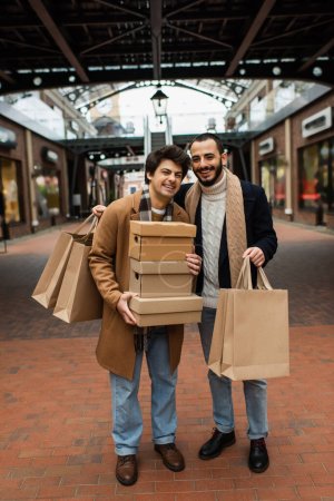 longitud completa de hombres homosexuales alegres con bolsas de compras y cajas de zapatos cerca del edificio con tiendas en el fondo