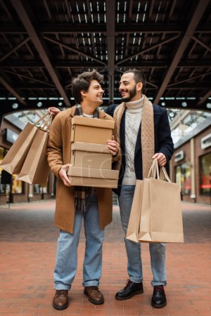 volle Länge der fröhlichen und stilvollen homosexuellen Männer mit Einkaufstaschen und Schuhkartons, die einander auf der städtischen Straße anschauen