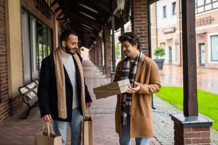 junger schwuler Mann schaut mit Einkaufstaschen auf Schuhkarton neben bärtigem Freund