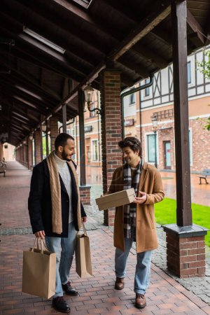 full length of happy gay man holding shoebox near bearded boyfriend with shopping bags on urban street