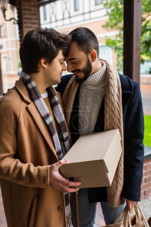 young gay man with closed eyes holding shoebox while standing face to face with happy bearded boyfriend 