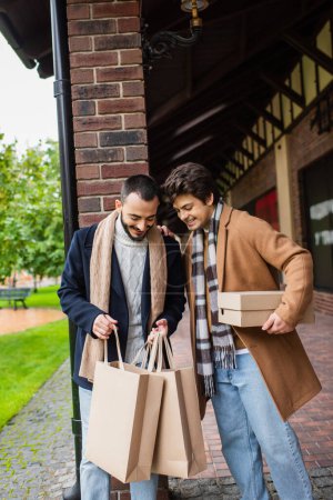 alegre gay los hombres en elegante ropa buscando en compras bolsas cerca de edificio en ciudad calle