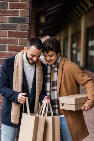 smiling homosexual couple in stylish outfit looking into shopping bags near brick column on city street