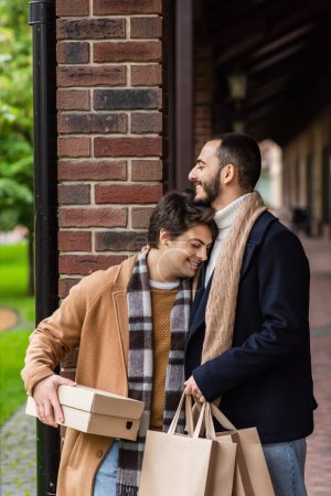 glücklicher schwuler Mann mit Zahnspange und geschlossenen Augen, der einen Schuhkarton hält, während er sich mit Einkaufstaschen auf einen bärtigen Freund stützt