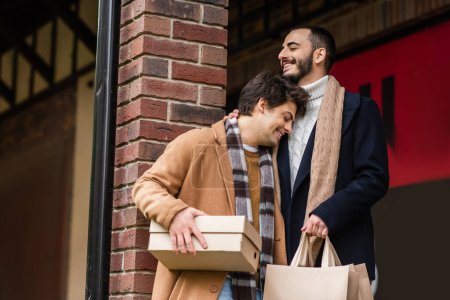 stylish and happy gay couple with purchases standing with closed eyes near brick column