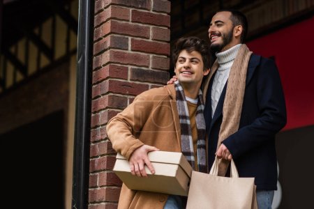 fröhliches und stylisches schwules Paar mit Einkaufstaschen und Schuhkarton, das in der Nähe der Ziegelsäule wegschaut