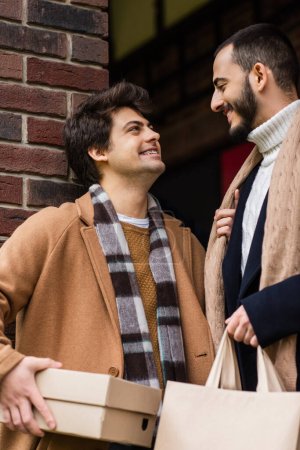 joyful and trendy gay couple with purchases smiling at each other near brick wall outdoors