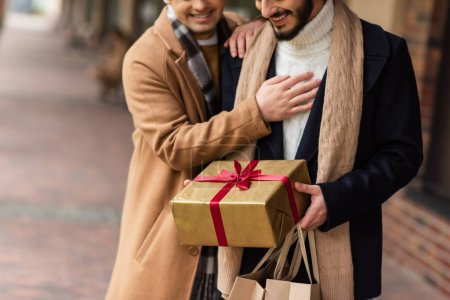 Freigestellter Blick auf bärtigen schwulen Mann mit Weihnachtsgeschenk und Einkaufstüten in der Nähe von Freund auf verschwommener Straße