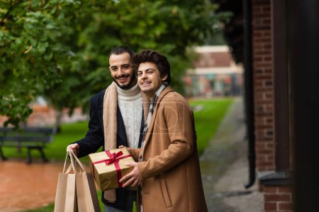 fröhliche und modische schwule Männer mit Weihnachtsgeschenk und Einkaufstüten, die draußen vor der Kamera stehen