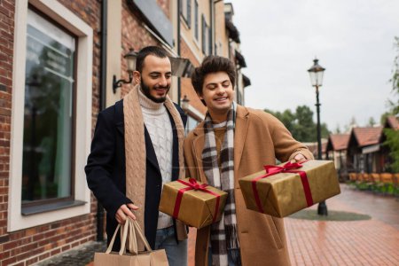 happy gay man holding gift boxes near bearded boyfriend with shopping bags and store on street