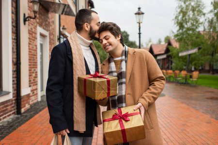 bearded gay man kissing head of happy boyfriend holding Christmas presents on city street