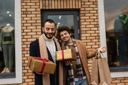 stylisches homosexuelles Paar hält Weihnachtsgeschenkboxen und Einkaufstüten in der Nähe des Geschäfts mit Vitrinen im Hintergrund