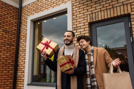 bärtiger schwuler Mann hält Weihnachtsgeschenkboxen in der Nähe von fröhlichem Freund und Geschäft mit Vitrinen