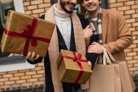 Ausgeschnittene Ansicht eines glücklichen und trendigen homosexuellen Paares mit Einkaufstaschen und Geschenkboxen auf der Straße der Stadt