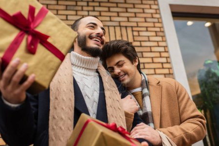 low angle view of bearded gay man holding gift boxes near happy boyfriend with closed eyes on street