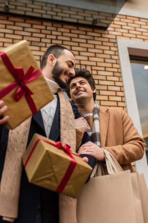 low angle view of cheerful gay men with Christmas presents and shopping bags near building on street
