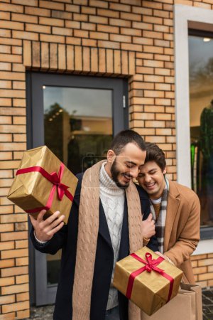 cheerful gay man laughing near bearded boyfriend holding Christmas gift boxes near shop on street