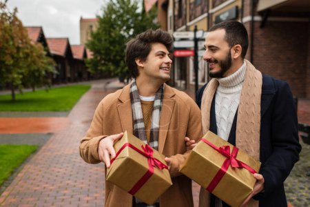 heureux gay couple dans branché manteaux et foulards tenant cadeaux boîtes et regarder l 'autre à l' extérieur