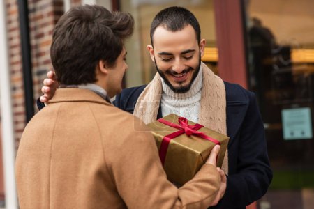 young man holding Christmas present near happy bearded gay partner on city street