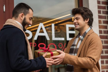 happy and stylish gay couple holding gift box near showcase with sale lettering