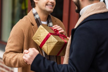 partial view of smiling gay couple holding hands near gift box on blurred street