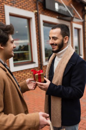 young gay man presenting small gift box to smiling bearded boyfriend near blurred shop on street