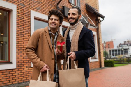 Photo for Cheerful and trendy gay men with Christmas present and shopping bags looking away near shop on urban street - Royalty Free Image