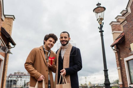 cheerful and stylish gay couple with shopping bags and small gift box looking at camera on city street
