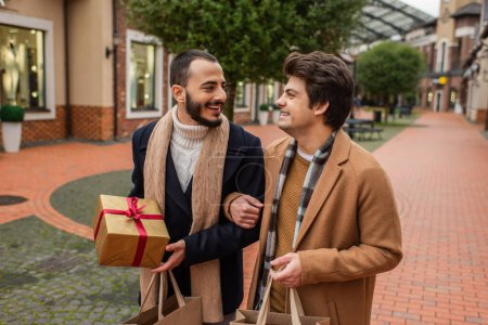 Téléchargez les photos : Heureux gay couple dans élégant vêtements tenue cadeau boîte et shopping sacs tout en souriant à l 'autre sur rue - en image libre de droit