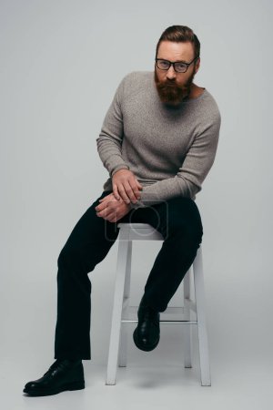 Full length of stylish bearded man in eyeglasses sitting on chair on grey background 